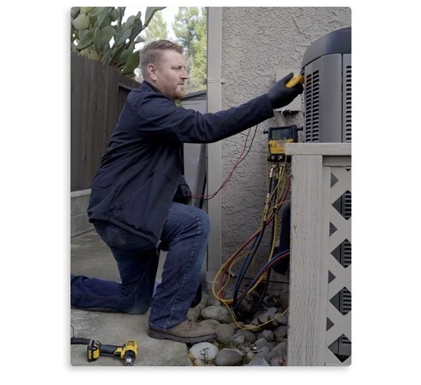 An HVAC technician kneels, inspecting an outdoor air conditioning unit